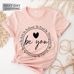 May include: A pink t-shirt with a black circle graphic that says "be you" with the words "Be Brave Be Bold Be Brilliant Be Kind Be Thoughtful Be Worthy Be Beautiful" circling around the graphic.