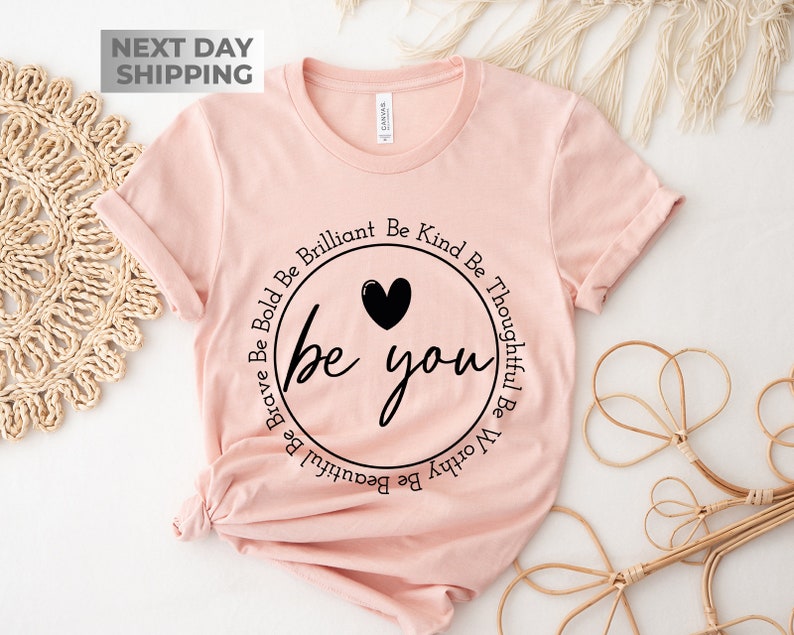 May include: A pink t-shirt with a black circle graphic that says "be you" with the words "Be Brave Be Bold Be Brilliant Be Kind Be Thoughtful Be Worthy Be Beautiful" circling around the graphic.
