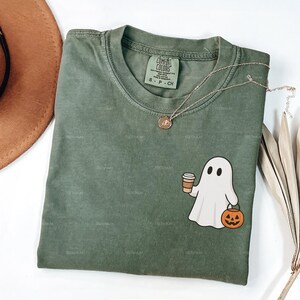 May include: A moss green Comfort Colors t-shirt featuring a cartoon ghost holding a coffee cup and a jack-o'-lantern. A gold necklace is draped across the shirt. The word "MOSS" is at the bottom.