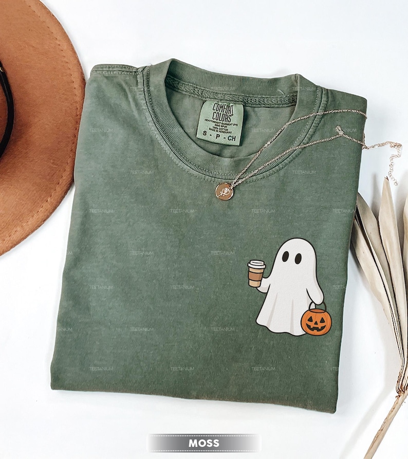 May include: A moss green Comfort Colors t-shirt featuring a cartoon ghost holding a coffee cup and a jack-o'-lantern. A gold necklace is draped across the shirt. The word "MOSS" is at the bottom.