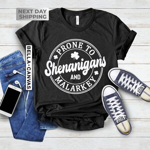 Op de afbeelding: Zwart T-shirt met een witte cirkelvormige afbeelding met de tekst "Prone to Shenanigans and Malarkey" met een klavertje vier in de cirkel.
