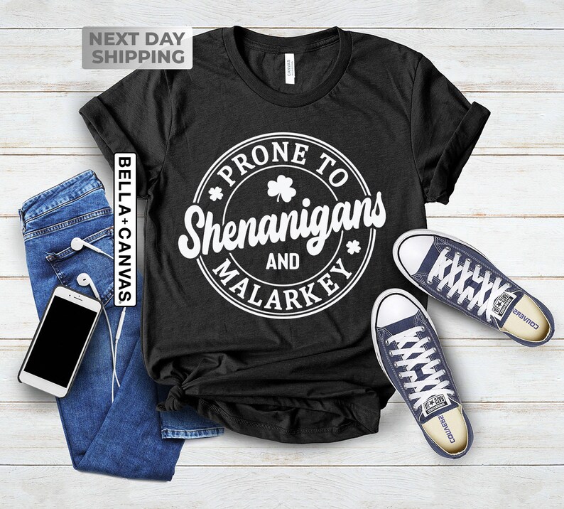 Op de afbeelding: Zwart T-shirt met een witte cirkelvormige afbeelding met de tekst "Prone to Shenanigans and Malarkey" met een klavertje vier in de cirkel.