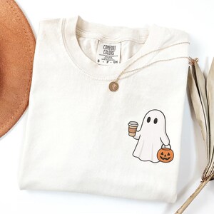 May include: Ivory t-shirt with a cartoon ghost holding a coffee cup and a jack-o'-lantern. The shirt has a crew neck and short sleeves. A gold necklace with a pendant is draped across the shirt.
