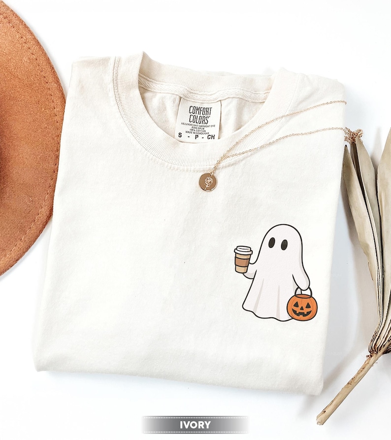 May include: Ivory t-shirt with a cartoon ghost holding a coffee cup and a jack-o'-lantern. The shirt has a crew neck and short sleeves. A gold necklace with a pendant is draped across the shirt.