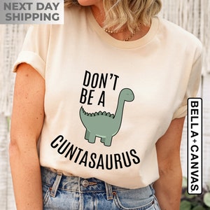 Pu&ograve; includere: Una t-shirt color crema con un'immagine di dinosauro verde e il testo "Don't be a Cuntasaurus".