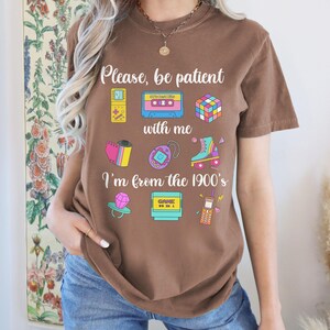 May include: A brown t-shirt featuring the phrase "Please, be patient with me, I'm from the 1900's" and illustrations of a cassette tape, Rubik's cube, roller skate, and other retro items. The shirt is worn with blue jeans.