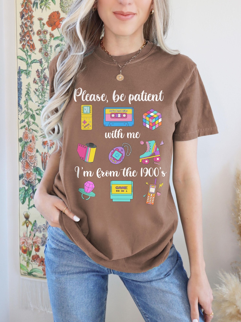 May include: A brown t-shirt featuring the phrase "Please, be patient with me, I'm from the 1900's" and illustrations of a cassette tape, Rubik's cube, roller skate, and other retro items. The shirt is worn with blue jeans.