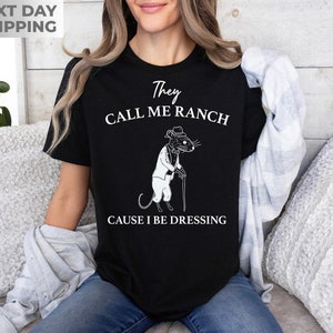 May include: Black t-shirt with a white graphic of a rat wearing a suit and holding a cane. The text on the shirt reads "They Call Me Ranch Cause I Be Dressing".