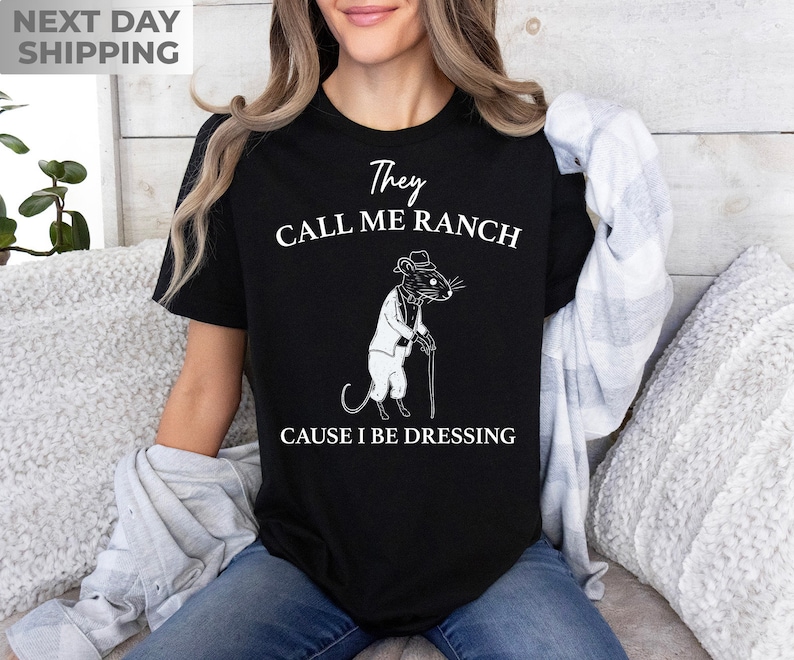 May include: Black t-shirt with a white graphic of a rat wearing a suit and holding a cane. The text on the shirt reads "They Call Me Ranch Cause I Be Dressing".