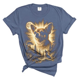 May include: Blue t-shirt featuring a detailed illustration of a Chihuahua dog with a radiant glow, set against a forest and moonlit sky. The design includes a smaller silhouette of a dog howling. The shirt is made of soft material.