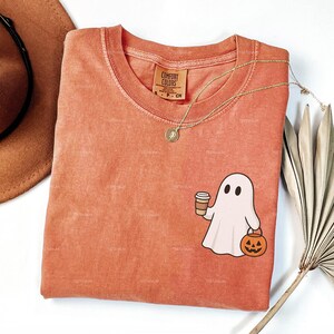 May include: Orange Comfort Colors t-shirt with a cartoon ghost holding a coffee cup and a jack-o'-lantern. A gold necklace and a brown hat are also visible. The shirt has a vintage, washed look, ideal for Halloween.