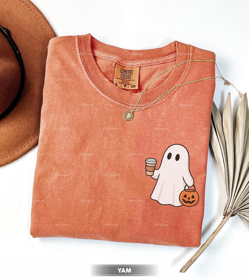 May include: Orange Comfort Colors t-shirt with a cartoon ghost holding a coffee cup and a jack-o'-lantern. A gold necklace and a brown hat are also visible. The shirt has a vintage, washed look, ideal for Halloween.