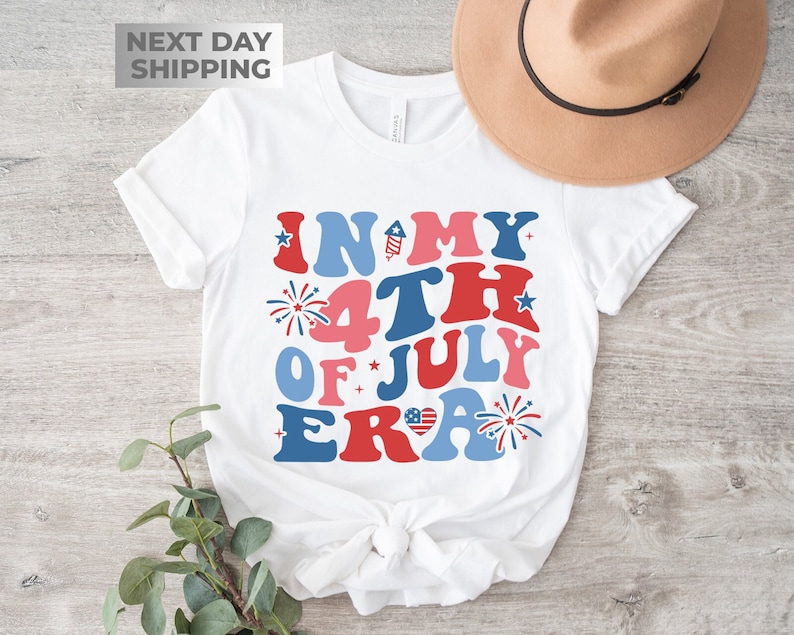 May include: A white t-shirt with the text 'In My 4th of July Era' in red, blue, and pink letters. The text is surrounded by stars and fireworks. The t-shirt is on a wooden surface with a brown hat on top of it.
