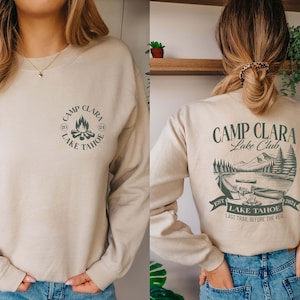 May include: A beige sweatshirt with a green graphic print on the front that says "Camp Clara Lake Tahoe" and a green graphic print on the back that says "Camp Clara Lake Club Est 2024 Lake Tahoe Last Trail Before The Veil"