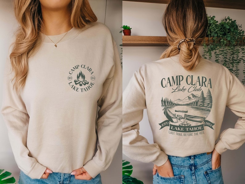 May include: A beige sweatshirt with a green graphic print on the front that says "Camp Clara Lake Tahoe" and a green graphic print on the back that says "Camp Clara Lake Club Est 2024 Lake Tahoe Last Trail Before The Veil"