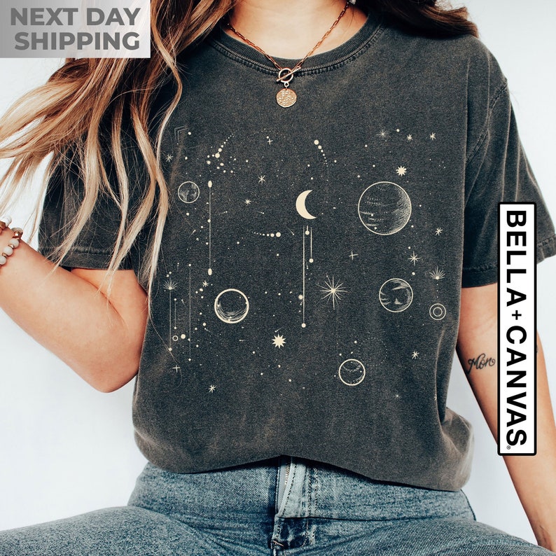 May include: A dark gray t-shirt with a celestial design featuring a crescent moon, stars, and planets in a light beige color. The text "BELLA + CANVAS" is printed on the right side of the shirt.