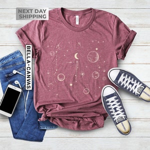 May include: A maroon t-shirt with a celestial design featuring a crescent moon, stars, and planets in a cream color. The text "Bella + Canvas" is printed on the left side of the shirt.