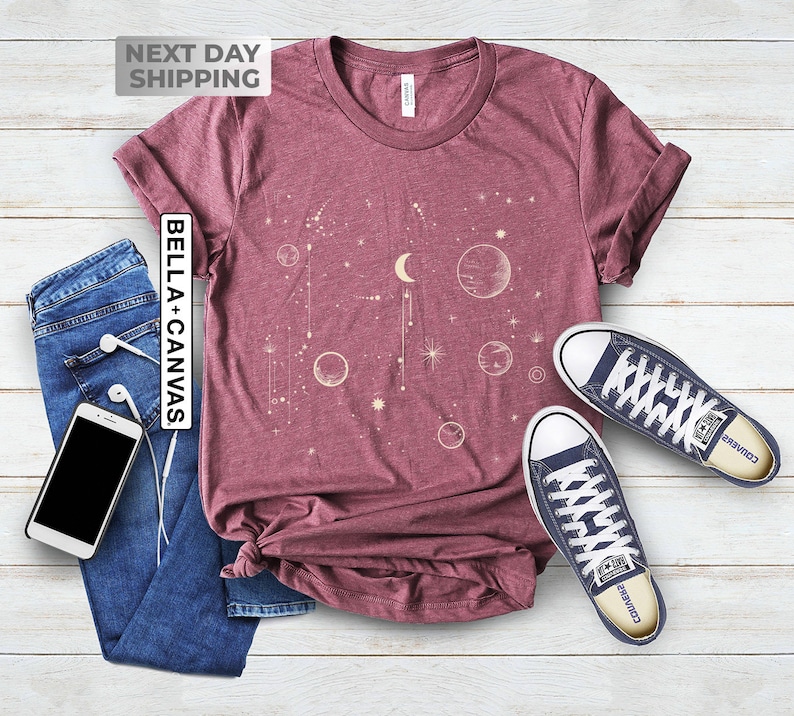 May include: A maroon t-shirt with a celestial design featuring a crescent moon, stars, and planets in a cream color. The text "Bella + Canvas" is printed on the left side of the shirt.