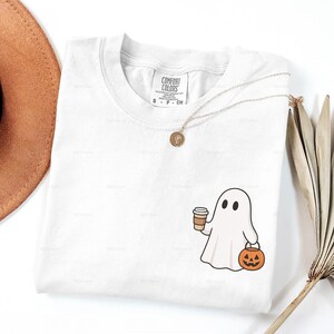 May include: White Comfort Colors t-shirt featuring a cartoon ghost holding a coffee cup and a jack-o'-lantern. A gold necklace with a pendant is draped on the shirt. The word "WHITE" is at the bottom.
