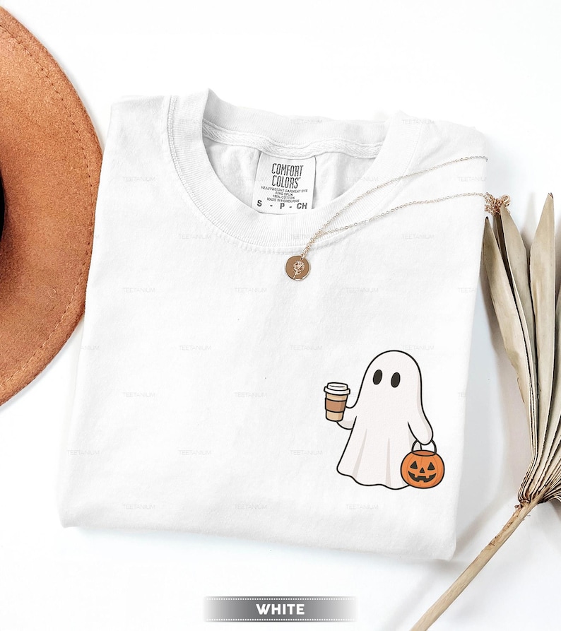 May include: White Comfort Colors t-shirt featuring a cartoon ghost holding a coffee cup and a jack-o'-lantern. A gold necklace with a pendant is draped on the shirt. The word "WHITE" is at the bottom.