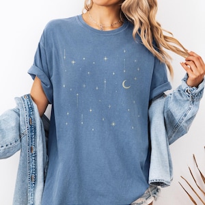 May include: Blue jean colored t-shirt with a celestial design featuring stars and a crescent moon. The shirt has a crew neck and short sleeves. The text "BLUE JEAN" is at the bottom of the image.