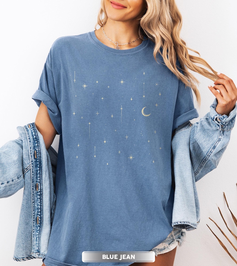 May include: Blue jean colored t-shirt with a celestial design featuring stars and a crescent moon. The shirt has a crew neck and short sleeves. The text "BLUE JEAN" is at the bottom of the image.