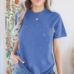 May include: A blue t-shirt with a celestial design featuring gold stars and a crescent moon. The shirt has short sleeves and a crew neck. The text "MYSTIC BLUE" is at the bottom of the image.