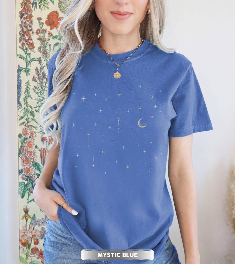 May include: A blue t-shirt with a celestial design featuring gold stars and a crescent moon. The shirt has short sleeves and a crew neck. The text "MYSTIC BLUE" is at the bottom of the image.