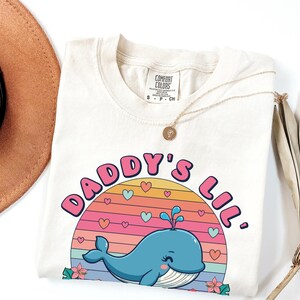 May include: A light beige t-shirt with a graphic design of a blue whale with pink hearts and a rainbow sunset. The text "Daddy's Lil'" is written in pink letters around the whale.