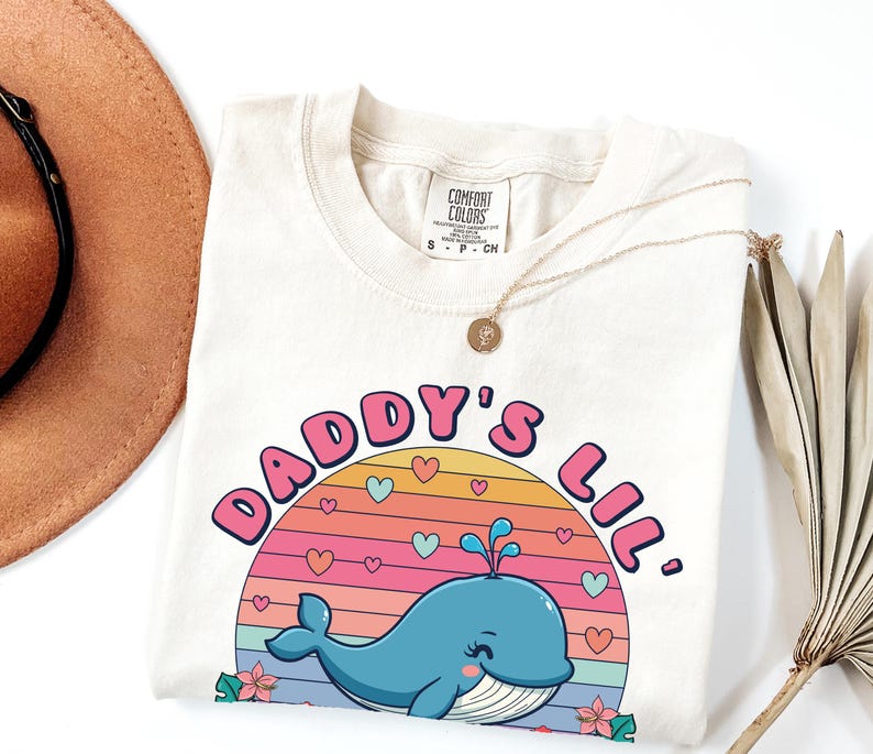 May include: A light beige t-shirt with a graphic design of a blue whale with pink hearts and a rainbow sunset. The text "Daddy's Lil'" is written in pink letters around the whale.