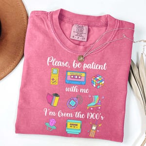 May include: A pink Comfort Colors t-shirt with the text "Please, be patient with me, I'm from the 1900's." The shirt features colorful graphics of a cassette tape, Rubik's cube, and other retro items. A silver necklace with a pendant is on the shirt.