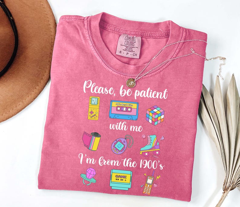 May include: A pink Comfort Colors t-shirt with the text "Please, be patient with me, I'm from the 1900's." The shirt features colorful graphics of a cassette tape, Rubik's cube, and other retro items. A silver necklace with a pendant is on the shirt.