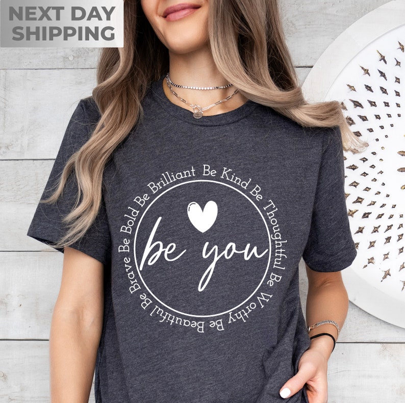 May include: A dark gray t-shirt with a white circle graphic that says "be you" with a heart in the center. The circle is surrounded by the words "Be Brave Be Bold Be Brilliant Be Kind Be Thoughtful Be Worthy Be Beautiful Be".