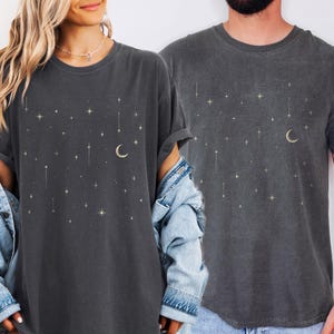 May include: Two dark gray t-shirts with a celestial design. The shirts feature a crescent moon and star pattern in a gold color. The t-shirts are made of a soft material and have a relaxed fit.
