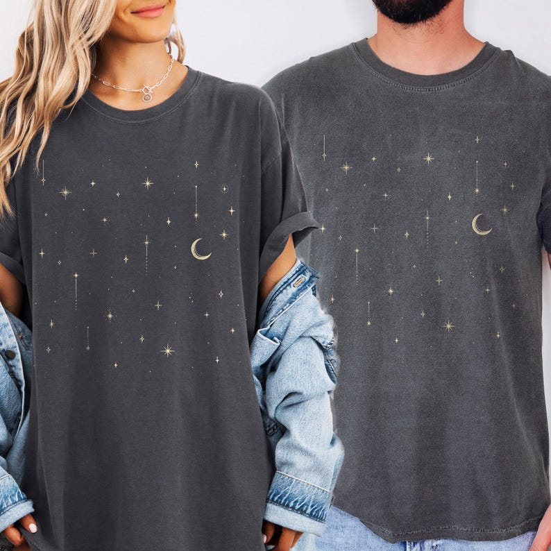 May include: Two dark gray t-shirts with a celestial design. The shirts feature a crescent moon and star pattern in a gold color. The t-shirts are made of a soft material and have a relaxed fit.