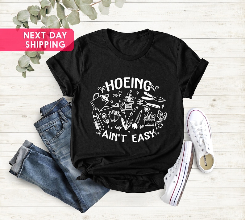 K&ouml;nnte beinhalten: Schwarzes T-Shirt mit wei&szlig;em Text "HOEING AIN'T EASY" und einer Grafik mit Gartenger&auml;ten und Pflanzen.