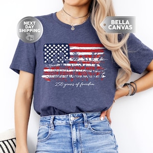 May include: Heather navy t-shirt featuring a distressed American flag design with red and white stripes and a blue field of stars. The text "250 years of freedom" is printed below the flag. The shirt is made of soft material.