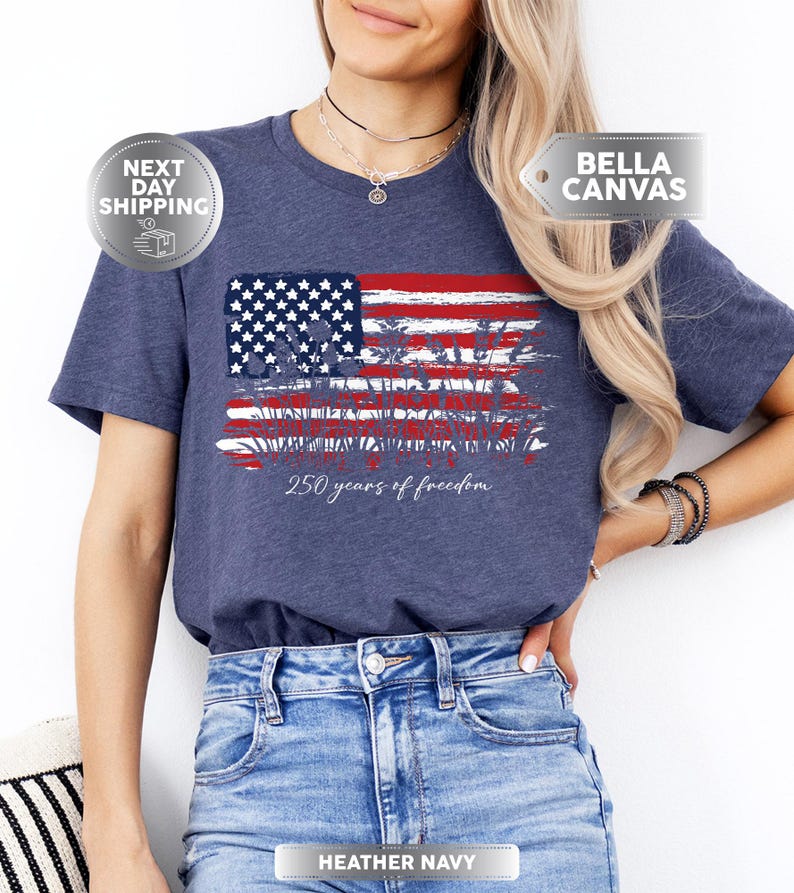 May include: Heather navy t-shirt featuring a distressed American flag design with red and white stripes and a blue field of stars. The text "250 years of freedom" is printed below the flag. The shirt is made of soft material.