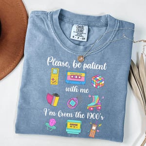 May include: A blue Comfort Colors t-shirt featuring the text "Please, be patient with me, I'm from the 1900's" and illustrations of a cassette tape, a Rubik's cube, and a roller skate. A silver necklace is draped across the shirt.