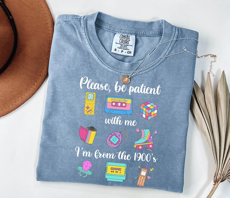 May include: A blue Comfort Colors t-shirt featuring the text "Please, be patient with me, I'm from the 1900's" and illustrations of a cassette tape, a Rubik's cube, and a roller skate. A silver necklace is draped across the shirt.