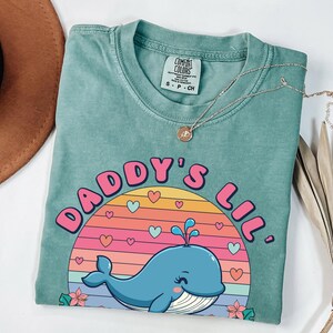 May include: A teal blue t-shirt with a graphic of a whale in a sunset with the text "Daddy's Lil'" in pink and white letters.