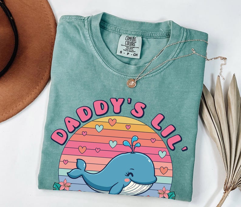 May include: A teal blue t-shirt with a graphic of a whale in a sunset with the text "Daddy's Lil'" in pink and white letters.
