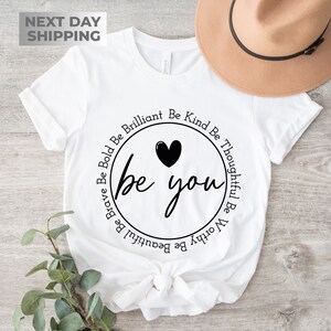 May include: White t-shirt with a black circle design that says "be you" with a heart above the text. The circle is surrounded by the words "Be Brave Be Bold Be Brilliant Be Kind Be Thoughtful Be Worthy Be Beautiful".