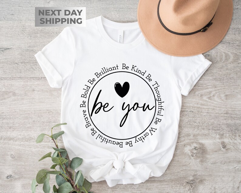 May include: White t-shirt with a black circle design that says "be you" with a heart above the text. The circle is surrounded by the words "Be Brave Be Bold Be Brilliant Be Kind Be Thoughtful Be Worthy Be Beautiful".