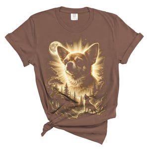 May include: Brown t-shirt featuring a detailed illustration of a Chihuahua dog with a radiant glow, a full moon, and a forest scene. The design includes a smaller dog silhouette. The shirt is knotted at the bottom.