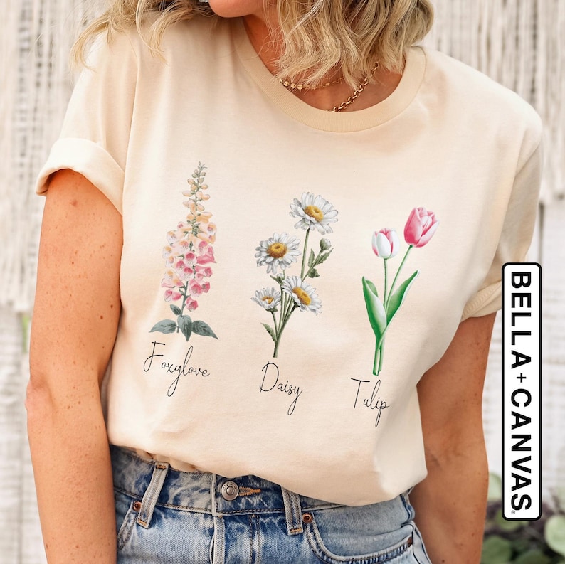 以下が含まれることがあります： クリーム色のTシャツ。フォックスグローブ、デイジー、チューリップの3つの花のイラストが描かれています。花には筆記体で名前が添えられています。Tシャツは半袖で、柔らかい素材で作られています。「BELLA+CANVAS」のブランド名が右側にプリントされています。