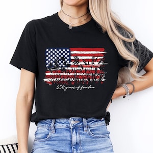 May include: Black t-shirt featuring a distressed American flag design with the text "250 years of freedom" in a script font. The flag design incorporates stars, stripes, and a silhouette of wheat. The shirt is paired with blue jeans.