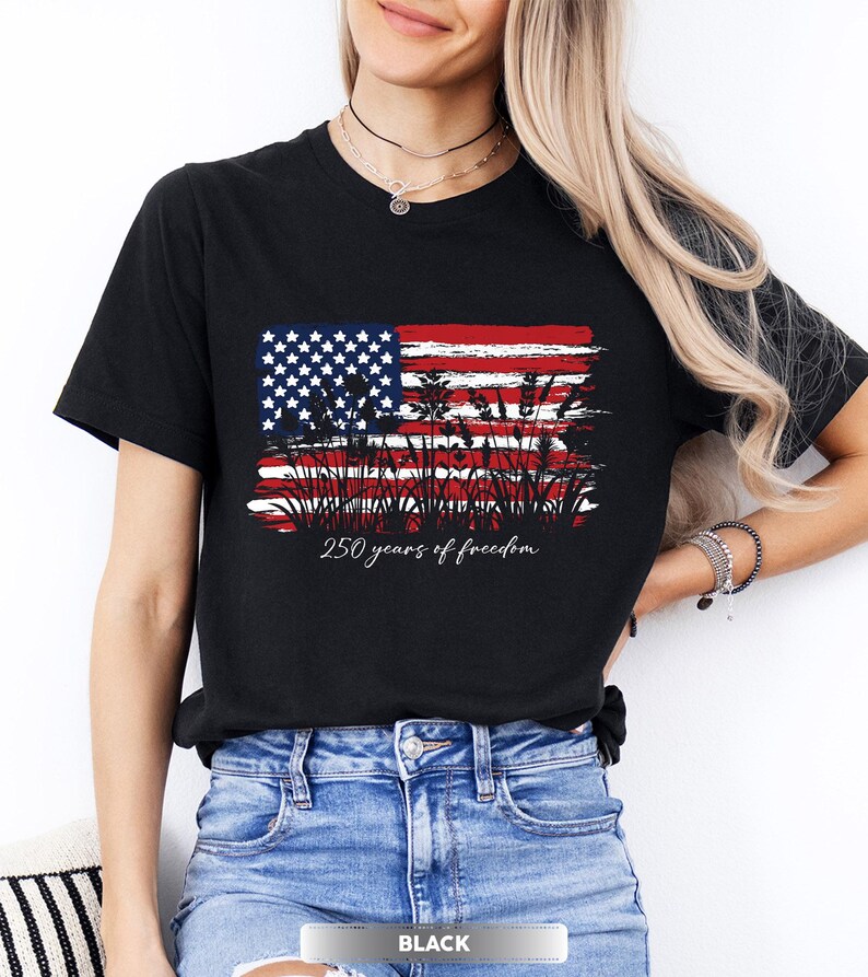 May include: Black t-shirt featuring a distressed American flag design with the text "250 years of freedom" in a script font. The flag design incorporates stars, stripes, and a silhouette of wheat. The shirt is paired with blue jeans.