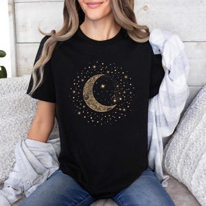 Może przedstawiać: Czarny t-shirt z złotym wzorem p&oacute;łksiężyca i gwiazd. Niebiańska grafika jest otoczona małymi gwiazdami. Koszulka ma klasyczny okrągły dekolt.