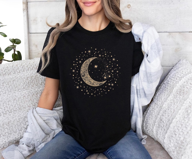 Może przedstawiać: Czarny t-shirt z złotym wzorem p&oacute;łksiężyca i gwiazd. Niebiańska grafika jest otoczona małymi gwiazdami. Koszulka ma klasyczny okrągły dekolt.